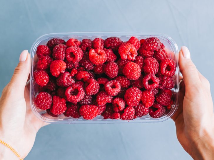 Fresh-ripe-raspberries-in-plastic-tub-on-light-blue-background